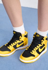 Fötter som bär svarta och gula Nike hög-top sneakers med vita strumpor, står på ljusblå strukturerad golv.