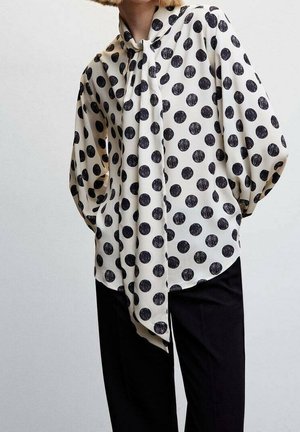 Personne portant un chemisier blanc à manches longues avec de gros pois noirs et une longue écharpe nouée autour du cou, associée à un pantalon noir.