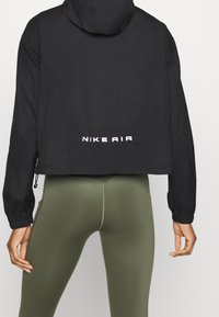 Veste noire courte avec capuche, offrant des poignets élastiques et un ourlet à cordon. Logo "NIKE AIR" en blanc au dos. Associée à des leggings vert olive.