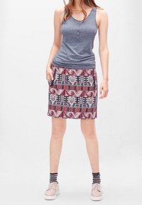 Kvinna som bär en ärmlös grå topp, mönstrad knä-lång kjol, randiga strumpor och ljusrosa snörskor, står med avslappnade händer.