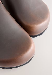 Braune Leder-Clogs mit abgerundeten Zehen und einer glatten, polierten Oberfläche. Strukturiert schwarze Gummisohlen und Nahtakzente entlang der Seiten.