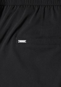 Black fabric waistband with an elastic band and a small rectangular metal label sewn near the stitching.