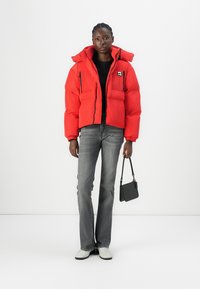 Red puffer jacket with a high collar, front pockets, and black zipper accents. Paired with gray flared jeans and white shoes, holding a small black bag.