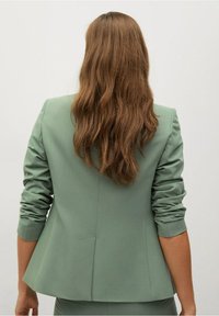 Jasnozielona marynarka o dopasowanym kroju z wyraźnymi ramionami i podwiniętymi rękawami. Gładki materiał z eleganckim krojem, z centralnym rozcięciem z tyłu.