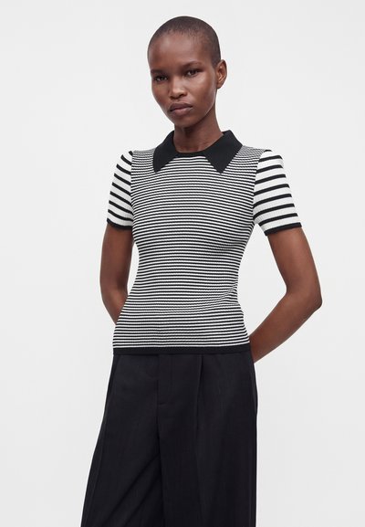 Femme aux cheveux courts portant un haut à manches courtes rayé noir et blanc avec un col noir et un pantalon noir, debout, les mains derrière le dos.