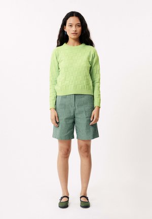 Lichtgroene, gestructureerde trui met een blokkenpatroon, gecombineerd met groene, gestreepte shorts. De outfit omvat groene schoenen met een band.