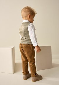 Un tout-petit portant un gilet gris clair sur une chemise blanche, un pantalon en velours côtelé brun et des chaussures beiges, se tenant à côté d'une forme géométrique blanche.