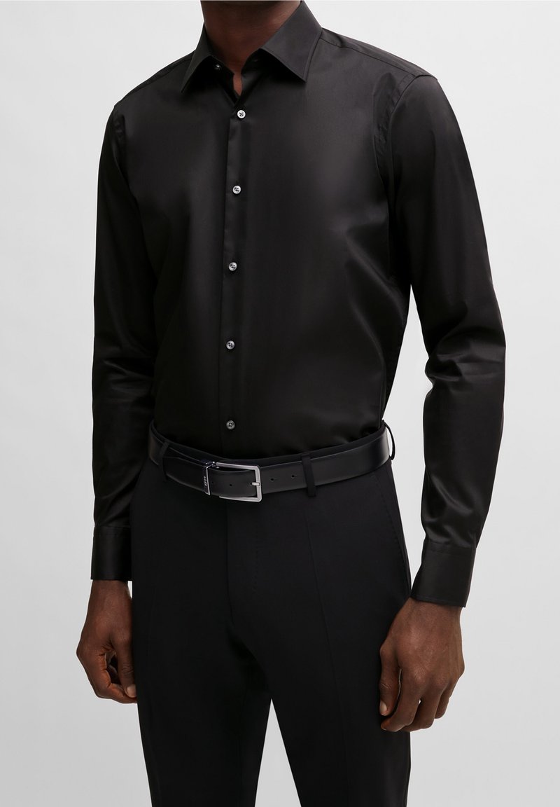 Homme portant une chemise noire cintrée à manches longues boutonnée, rentrée dans un pantalon noir avec une ceinture en cuir noire et une boucle argentée.