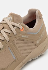 Beige Wanderschuh mit Obermaterial aus strukturiertem Stoff, auffälligem Gore-Tex-Logo und reflektierenden Akzenten. Ausgestattet mit Schnürsenkeln und Gummisohle.