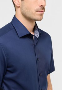 Marineblauw shirt met korte mouwen en knoopsluiting. Contrasterend geruite patroon op de binnenkraag. Glad, lichtgewicht materiaal met een getailleerde pasvorm.