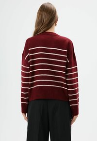 Bordeauxkleurige gebreide trui met horizontale crèmekleurige strepen, verlaagde schouders en geribde zoom, gecombineerd met zwarte broek. Achteraanzicht.