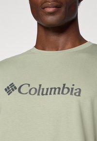 Světle zelené bavlněné tričko s kulatým výstřihem a černým logem "Columbia" na přední straně, nad abstraktním designem nášivky.