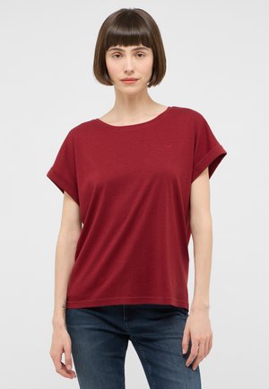 Woman with short brown hair wearing a loose red short-sleeve top and blue jeans, standing against a plain light background.