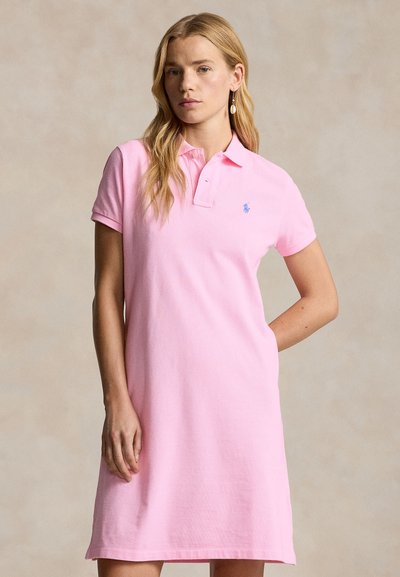 Femme portant une robe polo à manches courtes rose clair avec un petit logo bleu sur la poitrine, se tenant devant un fond beige uni.