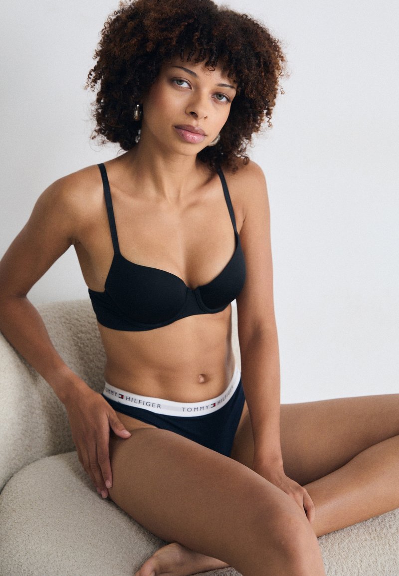 Soutien-gorge noir avec bretelles réglables et bonnets moulés associé à des culottes hipster noires, avec une ceinture blanche ornée du logo Tommy Hilfiger.