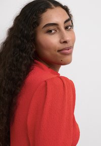 Blusa rossa con texture e maniche a sbuffo. La modella ha i capelli ricci, indossa trucco minimale e presenta una piccola voglia vicino al mento.