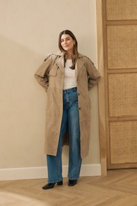 Femme debout contre un mur, portant un trench-coat beige, un haut blanc, un jean bleu et des bottes à talons noires dans un cadre intérieur minimaliste.