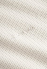 Biela texturovaná látka so vzorom mriežky, vybavená nenápadným vyšívaným logom, ktoré zdôrazňuje ľahkosť a priedušnosť materiálu.