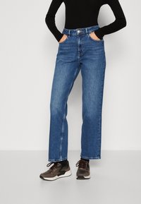 Osoba v modrých širokých džínách, s rukama v kapsách, černém dlouhém rukávu a hnědých teniskách, stojí proti bílému pozadí.