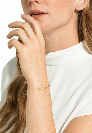 Goldene Armbandkette mit zwei grünen Akzentperlen, getragen an einem Handgelenk mit heller Hautfarbe, vor einem weißen hochgeschlossenen Kleidungsstück.