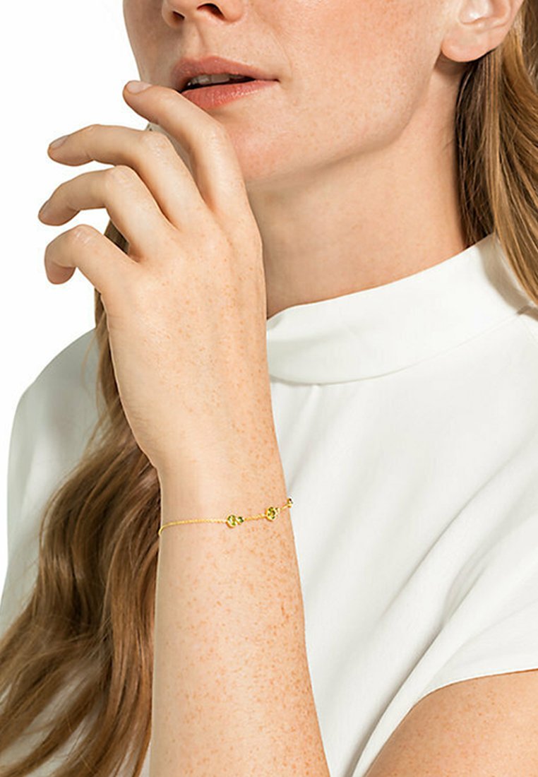 Gul länkarmband med två gröna accentpärlor, bärs på ett handleder med ljus hudton, mot en vit höghalshalsduk.