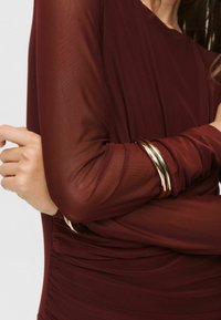 Bordeaux långärmad topp med genomskinliga ärmar och samlad textur, kompletterad med tre guldmetallarmband på handleden.