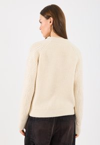 Neulottu kerma beige villapaita, jossa on tekstuurinen kuvio hihoissa, ribbaus helman ympärillä ja raglan-hihat, yhdistettynä tummiin korkeavyötäröisiin housuihin.