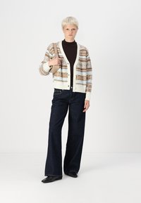 Cardigan en maille avec une base crème, des rayures marron et bleu, un col en V et des boutons. Associé à un jean large noir et un sac bandoulière beige.