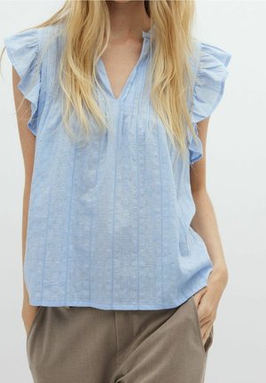 Frau mit langem blondem Haar, die eine hellblaue, ärmellose Bluse mit Rüschen und braune Hosen trägt, mit einer Hand in der Tasche vor weißem Hintergrund.