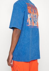 Blå grafisk t-shirt med en bleknad textur, med en orange tryck av hästar och stiliserad text på ryggen. Korta ärmar, avslappnad passform.