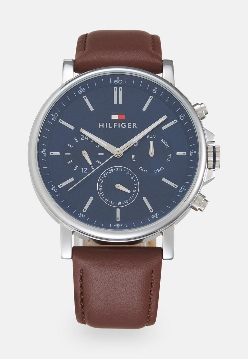 Montre en acier inoxydable argenté avec un cadran bleu marine, un bracelet en cuir marron, un affichage de la date et des sous-cadrans de chronographe. Marque HILFIGER présente.