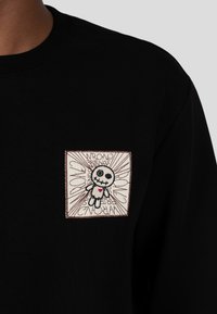 Felpa nera con una toppa ricamata a forma di quadrato che presenta una bambola in stile cartone animato, sfondo bianco, contorni neri e testo che dice "AMICI SCONTRATI".