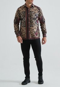 Chemise à boutons avec un motif paisley en noir, or et bordeaux. Manches longues, pantalon noir uni et chaussures noires complètent la tenue.