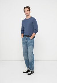 Cămașă albastră cu mâneci lungi, blugi evazați bleu deschis și mocasini negri cu accente albe. Modelul stă cu mâinile în buzunare, pe un fundal alb.