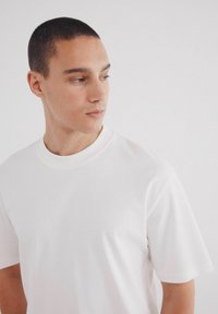 T-shirt bianca a maniche corte realizzata in tessuto morbido, con collo rotondo e un design semplice, caratterizzata da cuciture minime e una consistenza liscia.