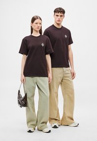 Dwie modelki noszą oversize'owe t-shirty, jedna w ciemnym brązie z diamentowym logo, druga w czarnym, zestawione z szerokimi spodniami w kolorze beżowym i zielonym.