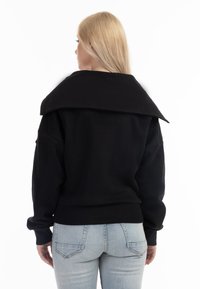Hanorac oversized negru, cu guler mare, manșete și tiv din material elastic. Fabrică moale, design minimalist, purtat cu blugi de un albastru deschis.