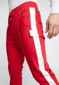 Nahaufnahme von roten Hosen mit elastischem Bund, vorderen Taschen, weißem Seitestreifen und einem kleinen weißen Markenetikett im Taschenbereich.