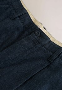 Dunkelblaue Hosen im Fischgrätmuster mit einem schwarzen Knopf und cremefarbenem Innenbund. Der Stoff weist eine glatte Textur auf und hat Falten.