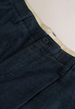 Dunkelblaue Hosen im Fischgrätmuster mit einem schwarzen Knopf und cremefarbenem Innenbund. Der Stoff weist eine glatte Textur auf und hat Falten.