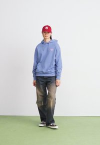 Light blue hoodie with front pocket and logo, paired with distressed dark jeans. Red cap and black sneakers complete the outfit.