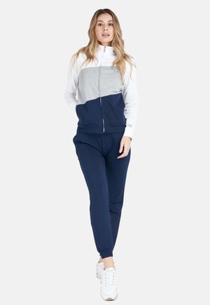 Une femme porte une veste de survêtement zippée blanche, grise et bleu marine, assortie à un pantalon de jogging bleu marine et à des baskets blanches, sur un fond uni.