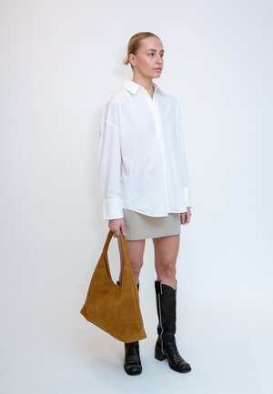 Femme portant une chemise blanche oversize boutonnée, une mini-jupe beige, des bottes noires montantes, tenant un grand sac à main en daim marron, debout devant un fond blanc.