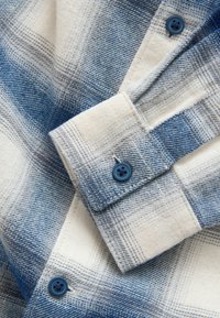 Blaues und weißes kariertes Flanellhemd mit Nahaufnahme einer geknöpften Manschette. Verfügt über kontrastierende blaue Knöpfe und eine weiche Textur.