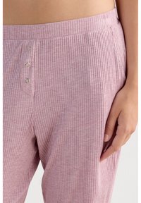 Close-up van een persoon die roze geribbelde broek draagt met drie metalen drukknoopjes en een zijzak, hand rustend bij de dij.