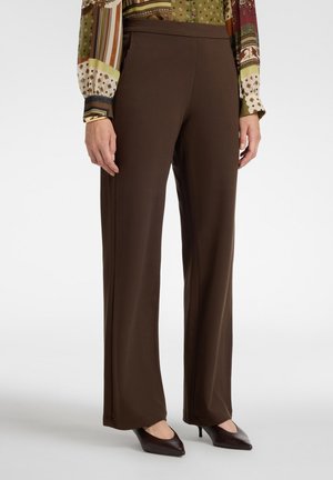 Donna che indossa pantaloni larghi marrone scuro, una camicetta a maniche lunghe con motivi e décolleté scure con punta appuntita, in piedi su sfondo chiaro.