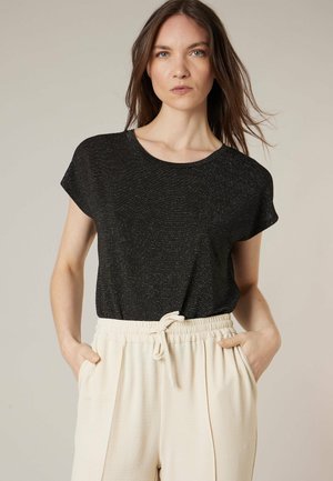 Femme aux cheveux bruns lâches portant un haut noir à manches courtes et un pantalon crème à cordon, debout les mains dans les poches devant un fond beige.