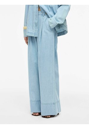 Személy világoskék, bőszárú farmernadrágot és hozzáillő túlméretezett farmerdzsekit visel, keze részben a zsebében, fekete nyitott orrú magassarkúval.