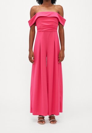 Kvinde iført en klar pink off-shoulder jumpsuit med vidde ben, plisseret front og sølvfarvede åbne sandaler på hvid baggrund.