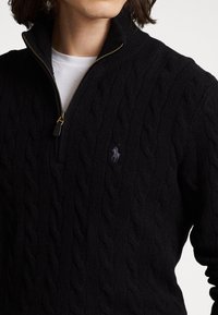 Schwarzer Strickpullover mit halber Reißverschlusskragen, gerippten Bündchen und einem dezenten Logo-Detail auf der Brust. Weiche Textur, warmes Material.